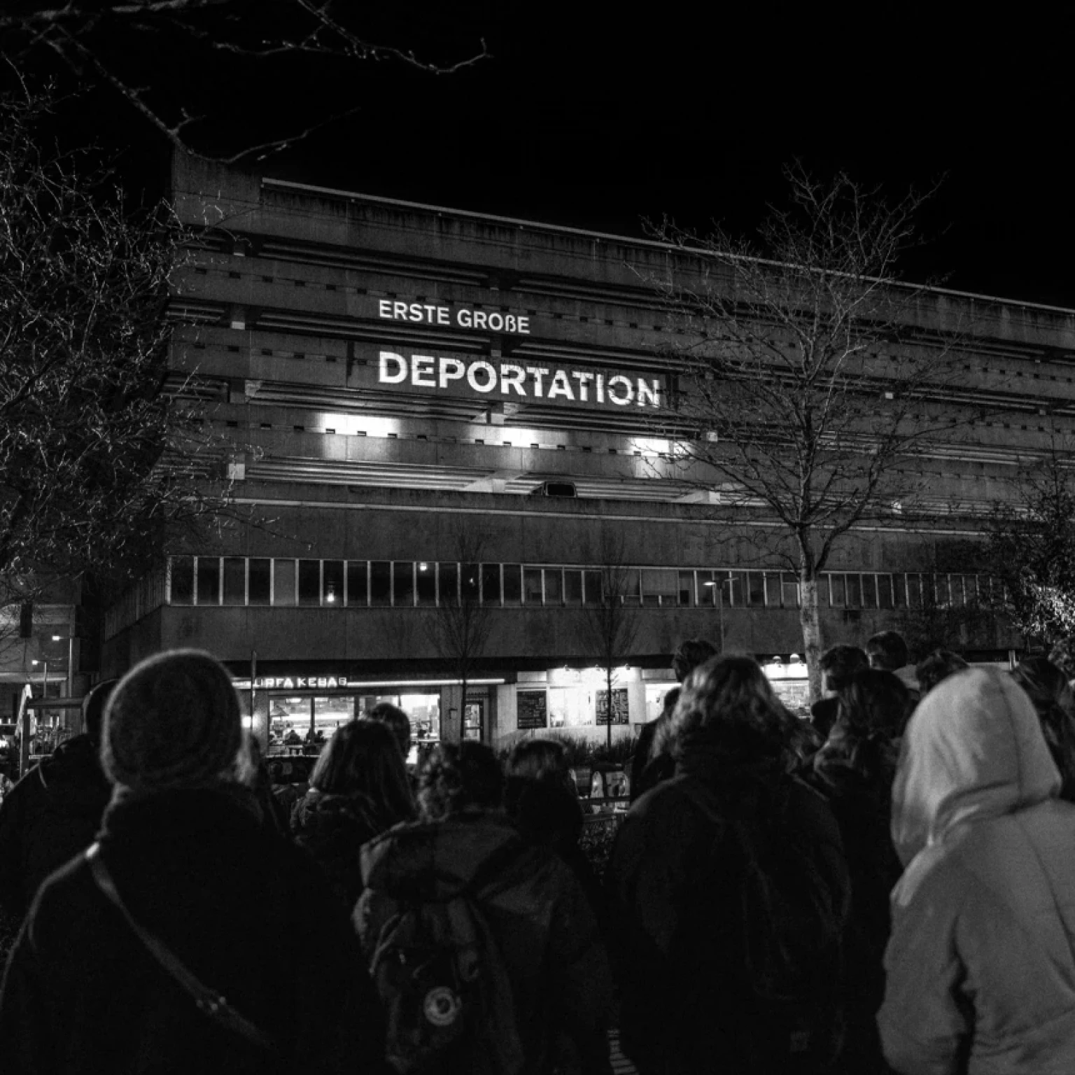 In a black-and-white shot, people can be seen standing in front of a façade projection with the words "First major deportation".